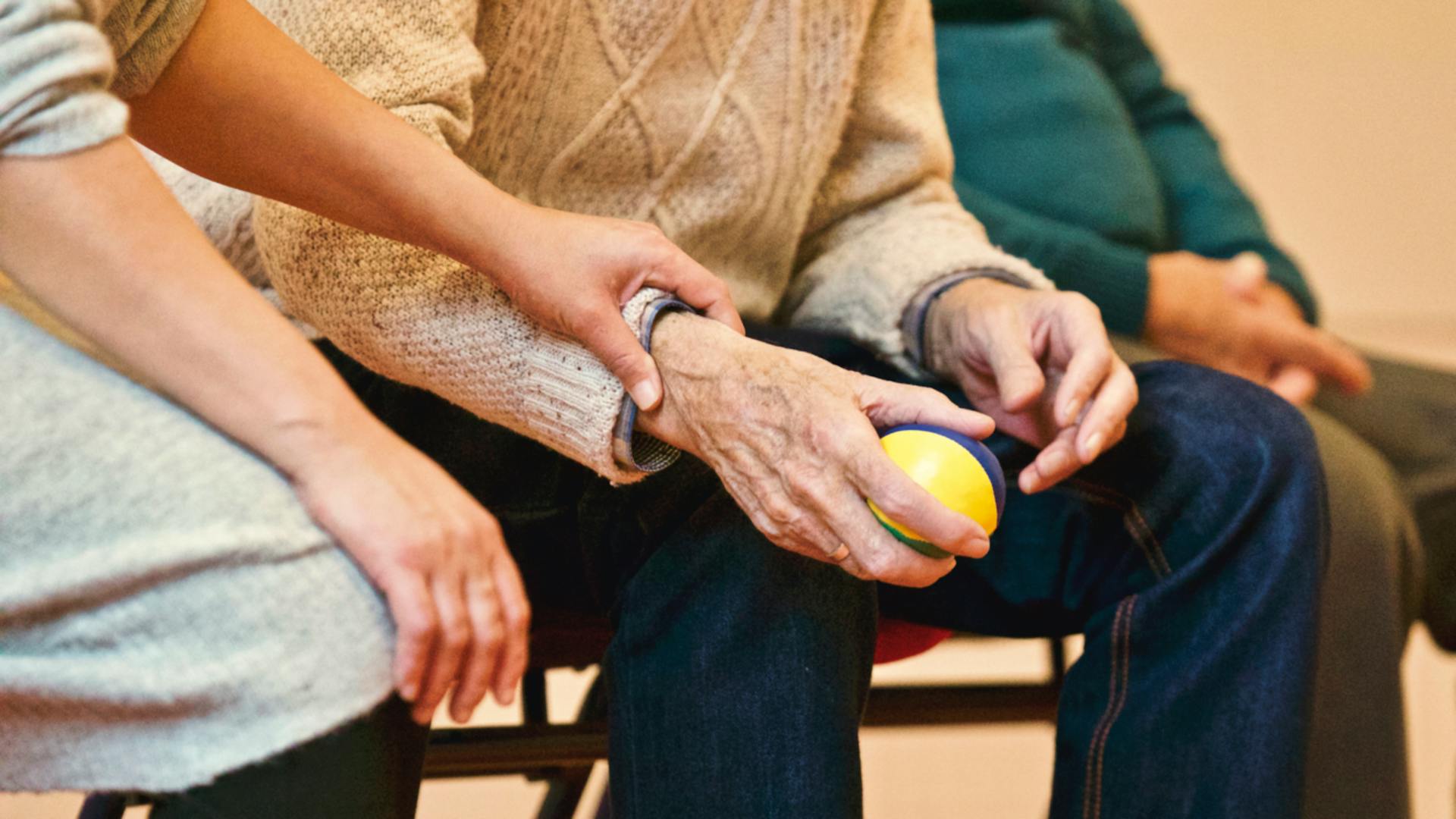 Young caregiver’s hand gently supporting an elderly hand holding a therapy ball